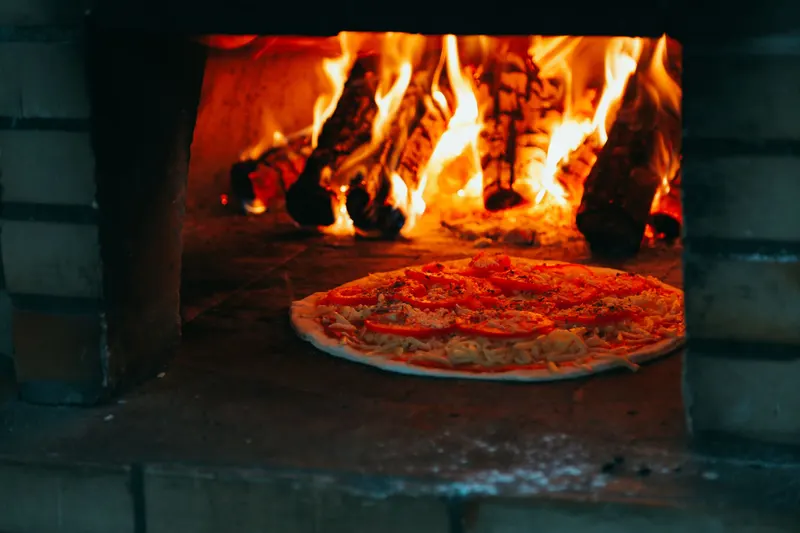 Holzfeuer im gemauerten Steinofen, eine Pizza backt davor